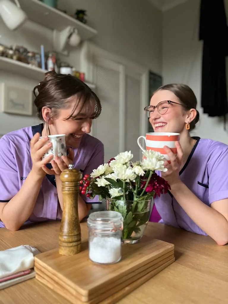 Two Quality Care Scotland carers in lilac uniforms sharing a laugh over a cup of tea
